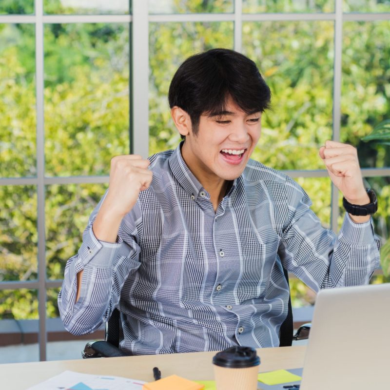 Yes! Happy excited Asian man raising his arm up to celebrate celebrating success. Young businessman using laptop computer at office desk he glad to receive good winner profits from the job